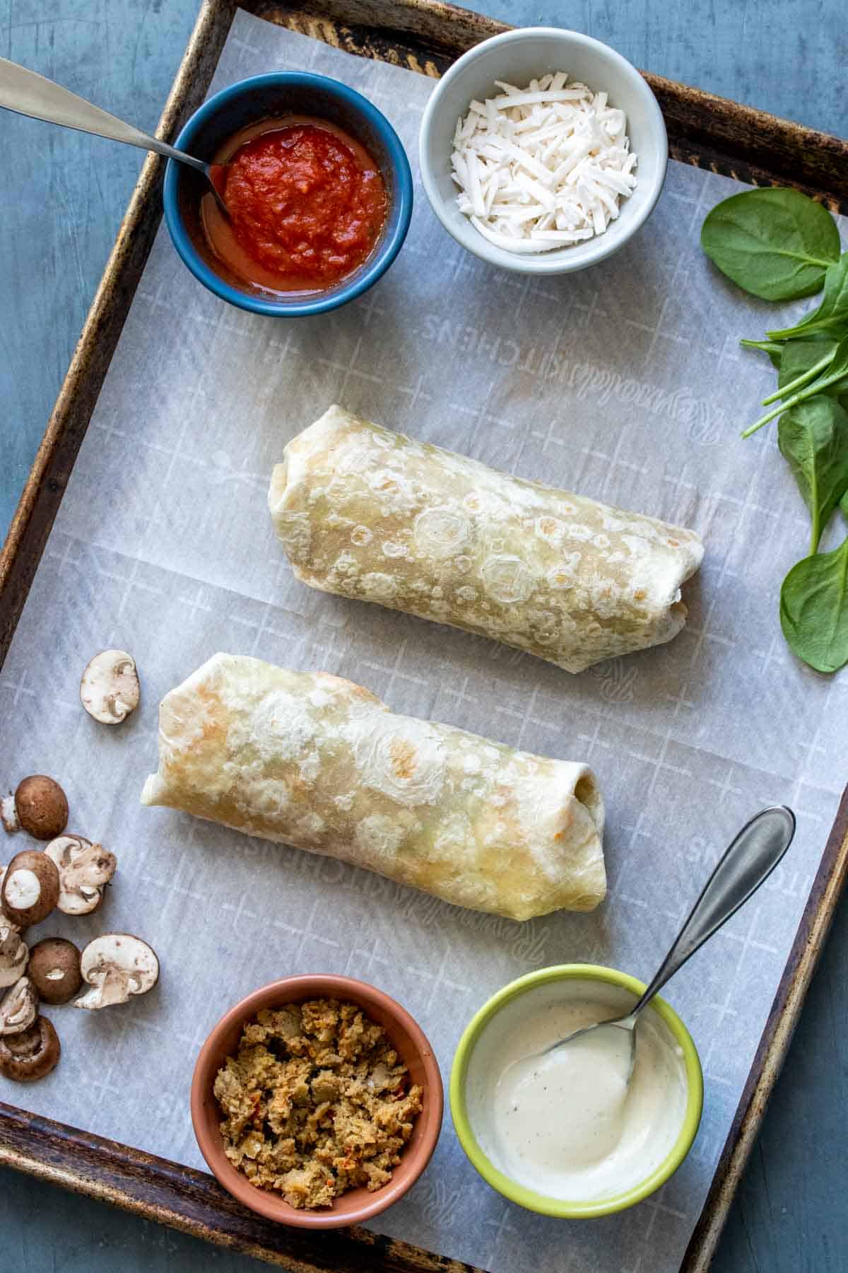 Two burritos on a parchment lined baking sheet with pizza ingredients around it