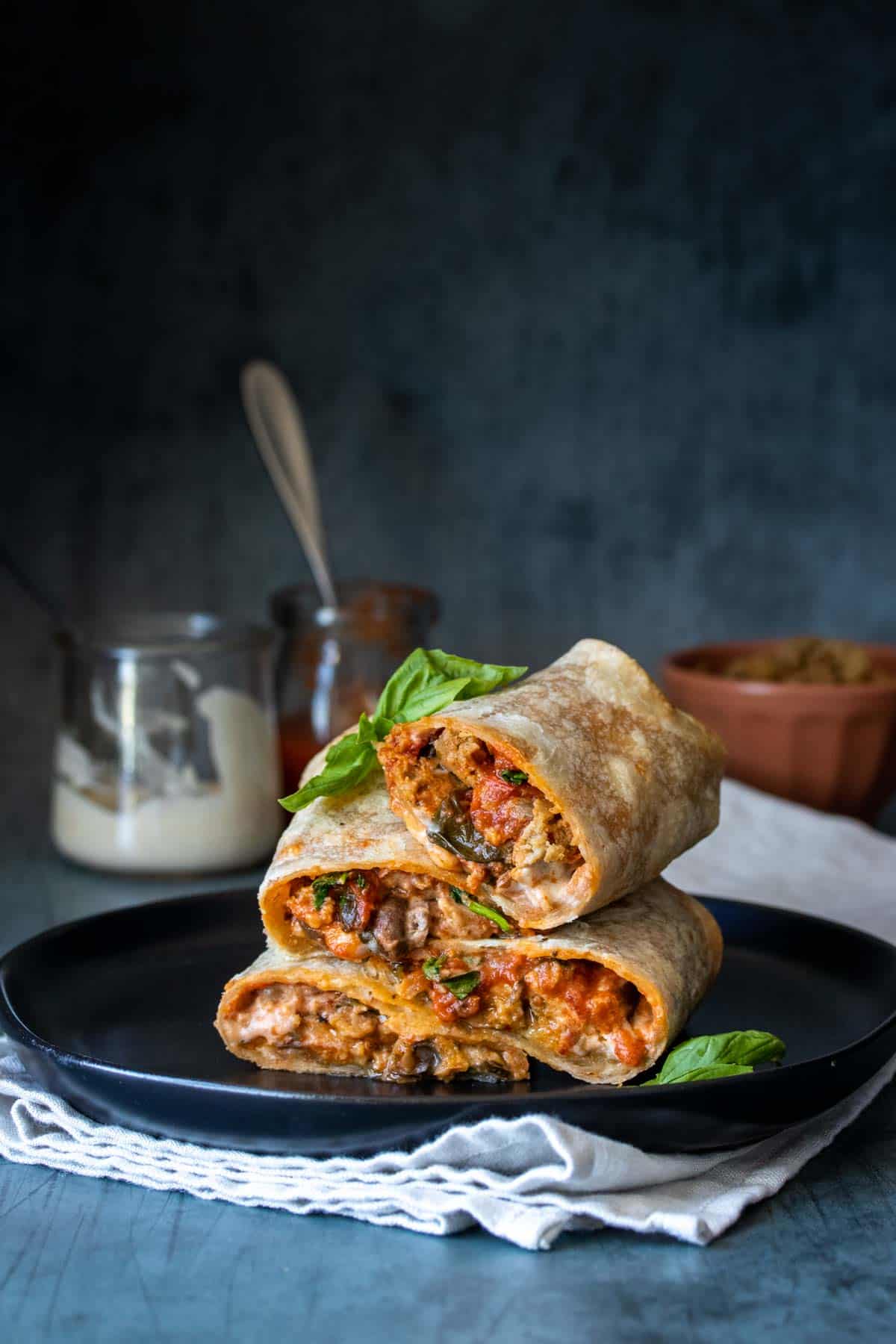 Pizza stuffed burritos cut in half and piled onto a black plate sitting on a folded towel on a grey surface and background