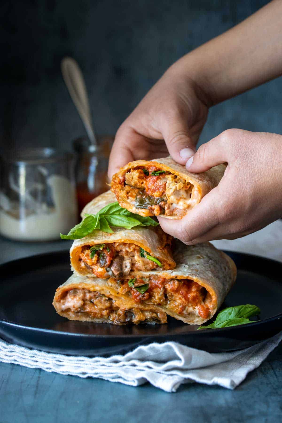 Two hand picking up half of a pizza themed burrito from a pile of them on a black plate with basil on it