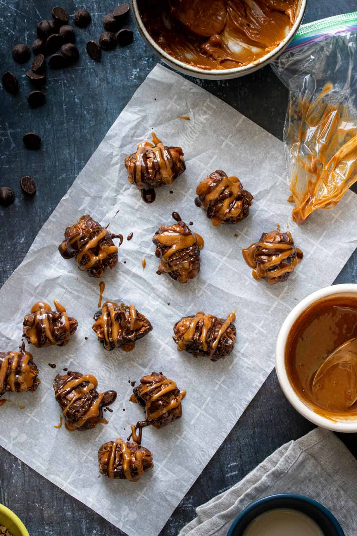 A group of dates on a piece of parchment paper drizzled with chocolate and caramel next to bowls of those