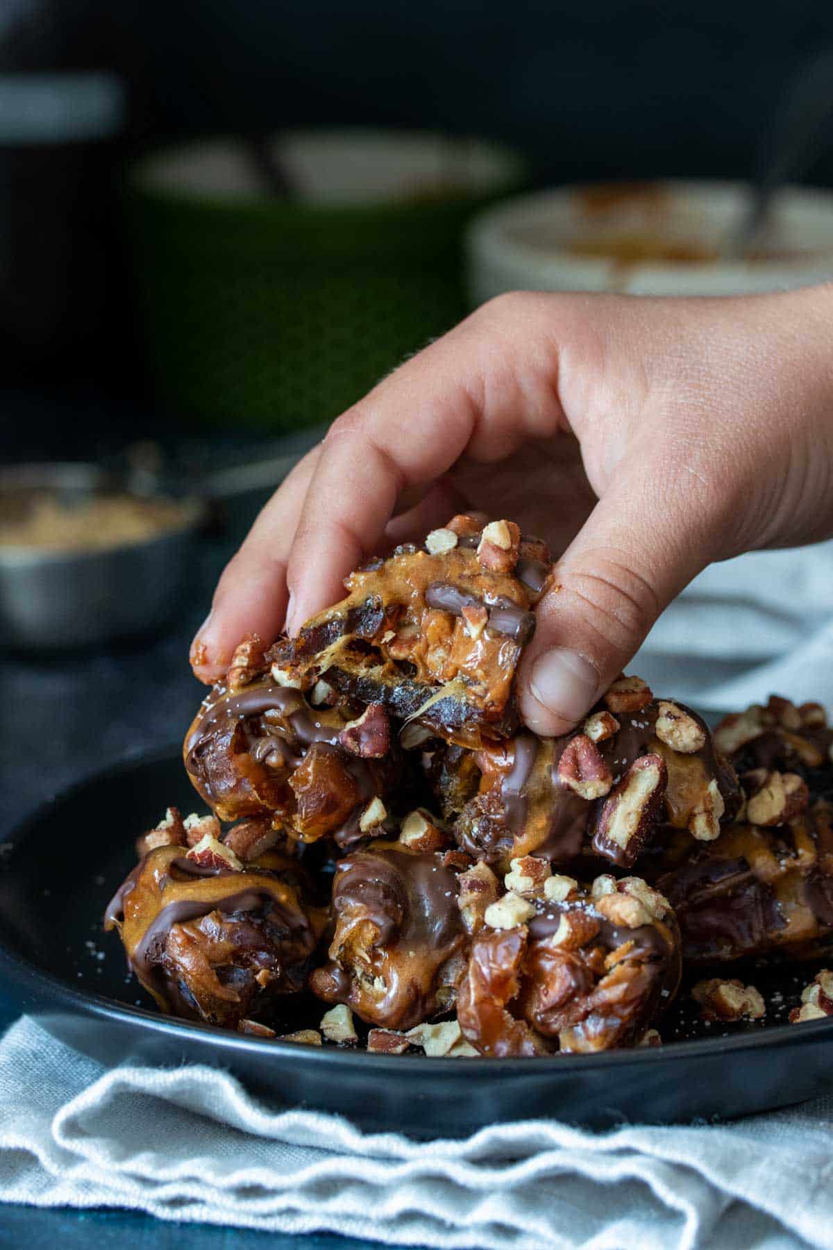 A hand grabbing a stuffed date covered in chocolate and caramel from a pile of them on a black plate