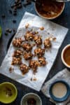 Top view of a group of chocolate and caramel covered stuffed dates on a piece of parchment laying on black plate next to bowls of ingredients