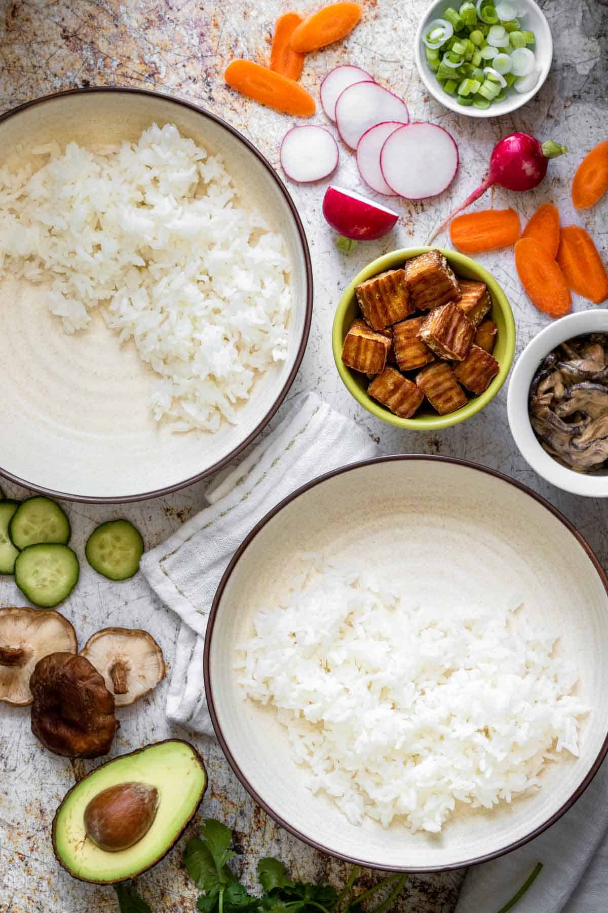 Overhead view of cream bowls with a brown rim filled with rice surrounded by cut veggies and tofu