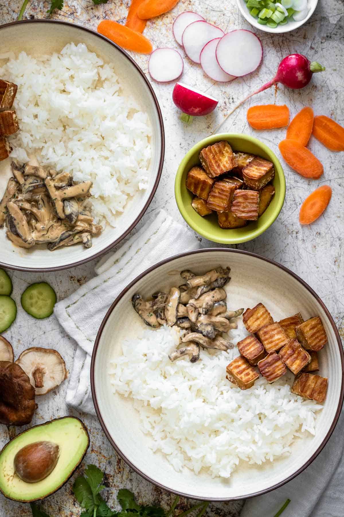 Rice, tofu and mushrooms in two cream bowls surrounded by more ingredients for sushi bowls