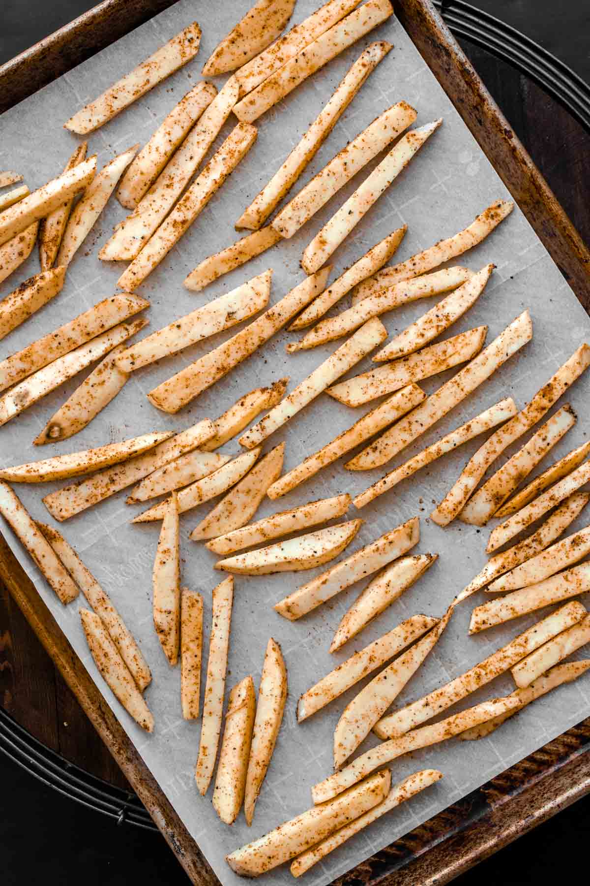 Cut sweet potatoes covered in seasoning on a piece of parchment paper on a baking sheet.