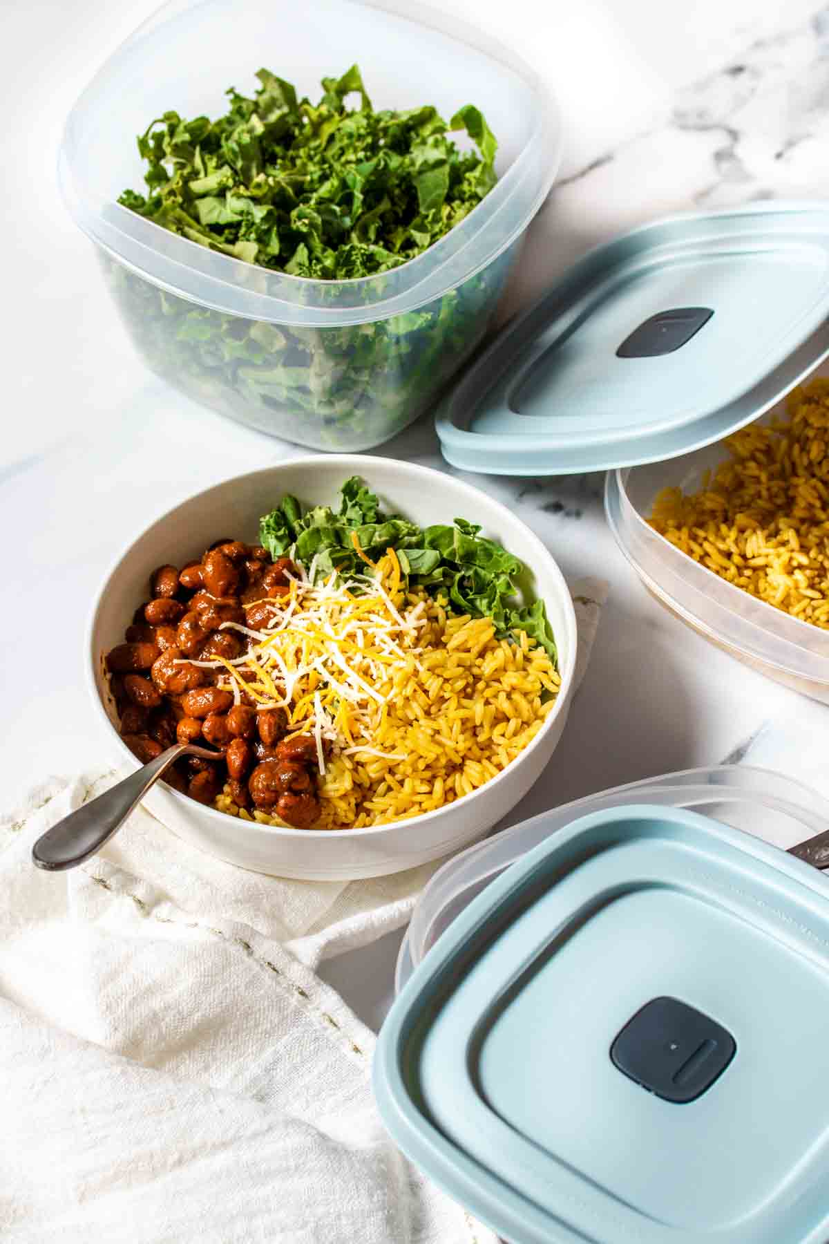 A white bowl with beans, rice and greens inside next to container with blue lids with those items in them.