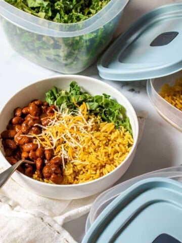 Beans, rice and greens in a white bowl with a spoon next to three containers with blue lids and food inside.