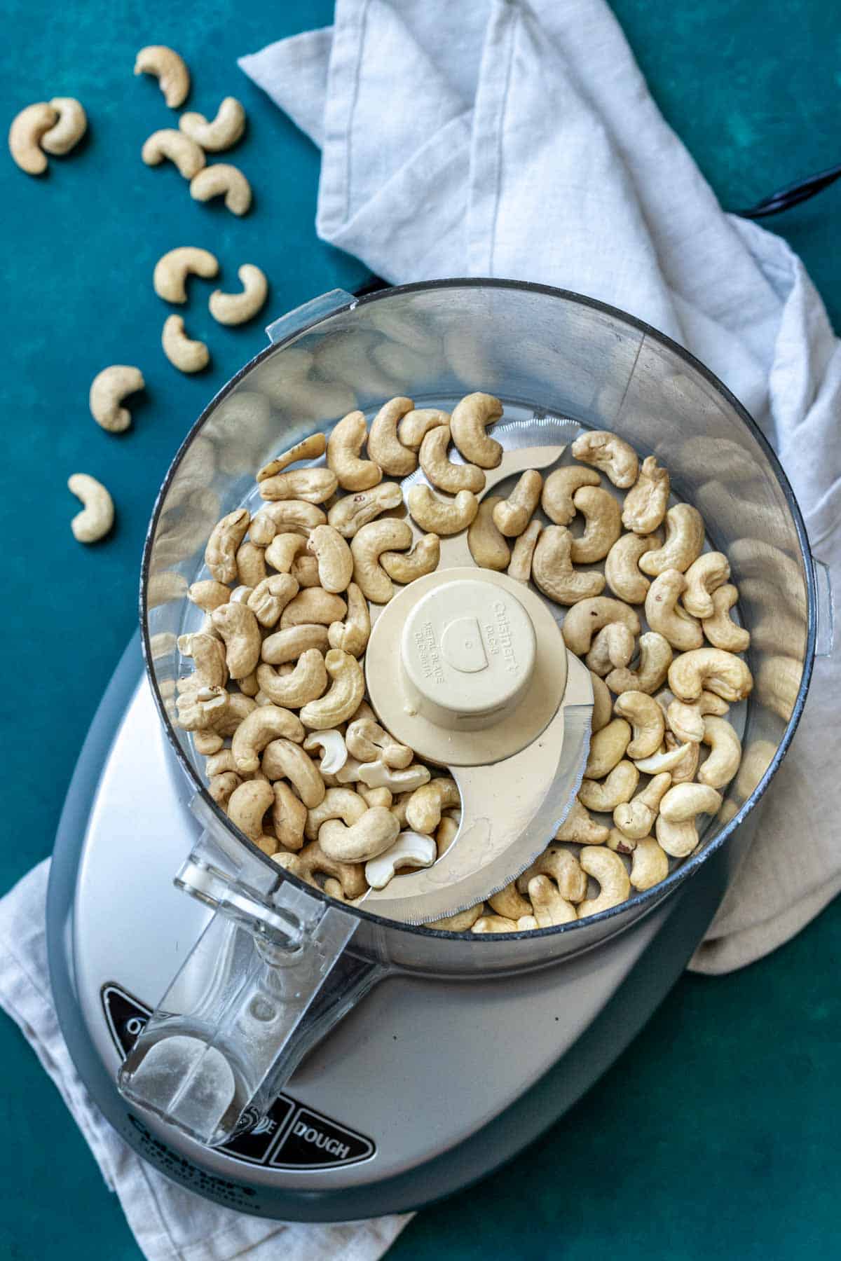 Top of a food processor with whole cashews inside sitting on a white towel.