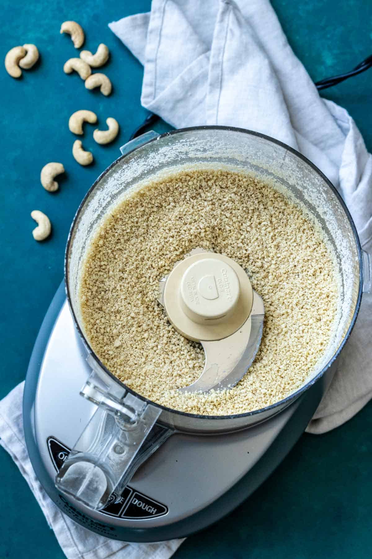 Top view of a food processor with blended cashews in it sitting on a towel on a dark turquoise surface.