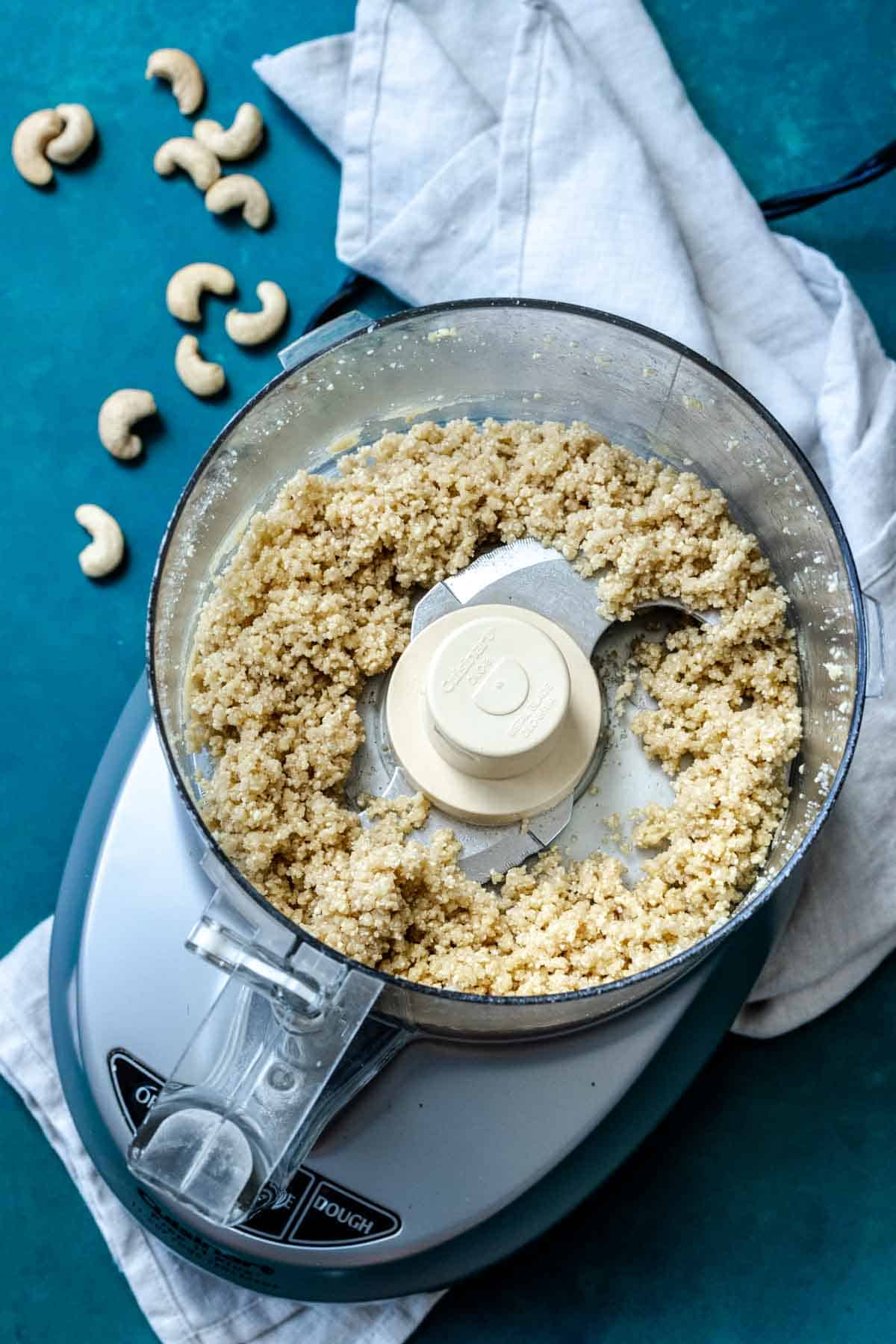 A blender on a white towel filled with a wet crumble of cashews and whole cashews on laying next to it.