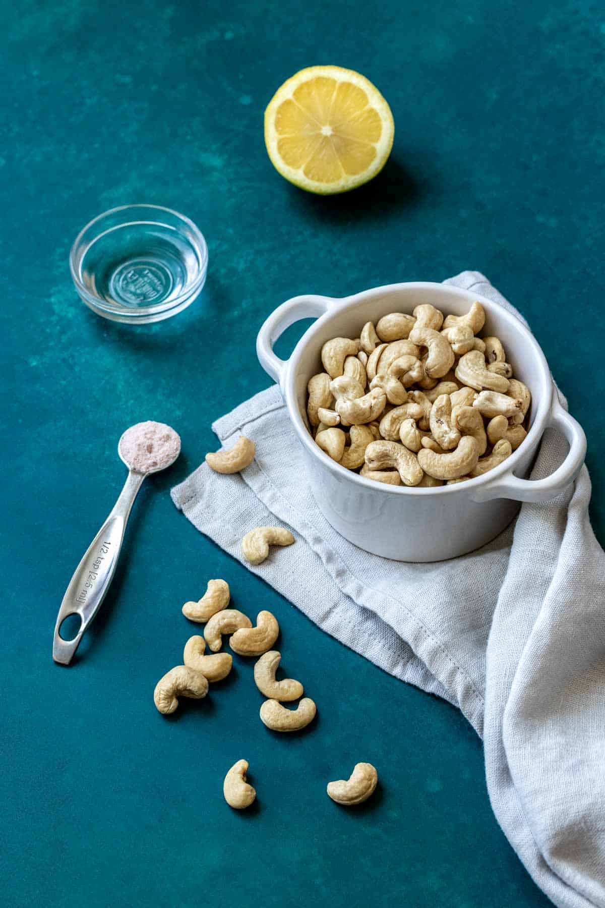 A dark turquoise backdrop with a bowl of cashews, half a lemon, a spoon of salt and a small bowl with lemon juice.