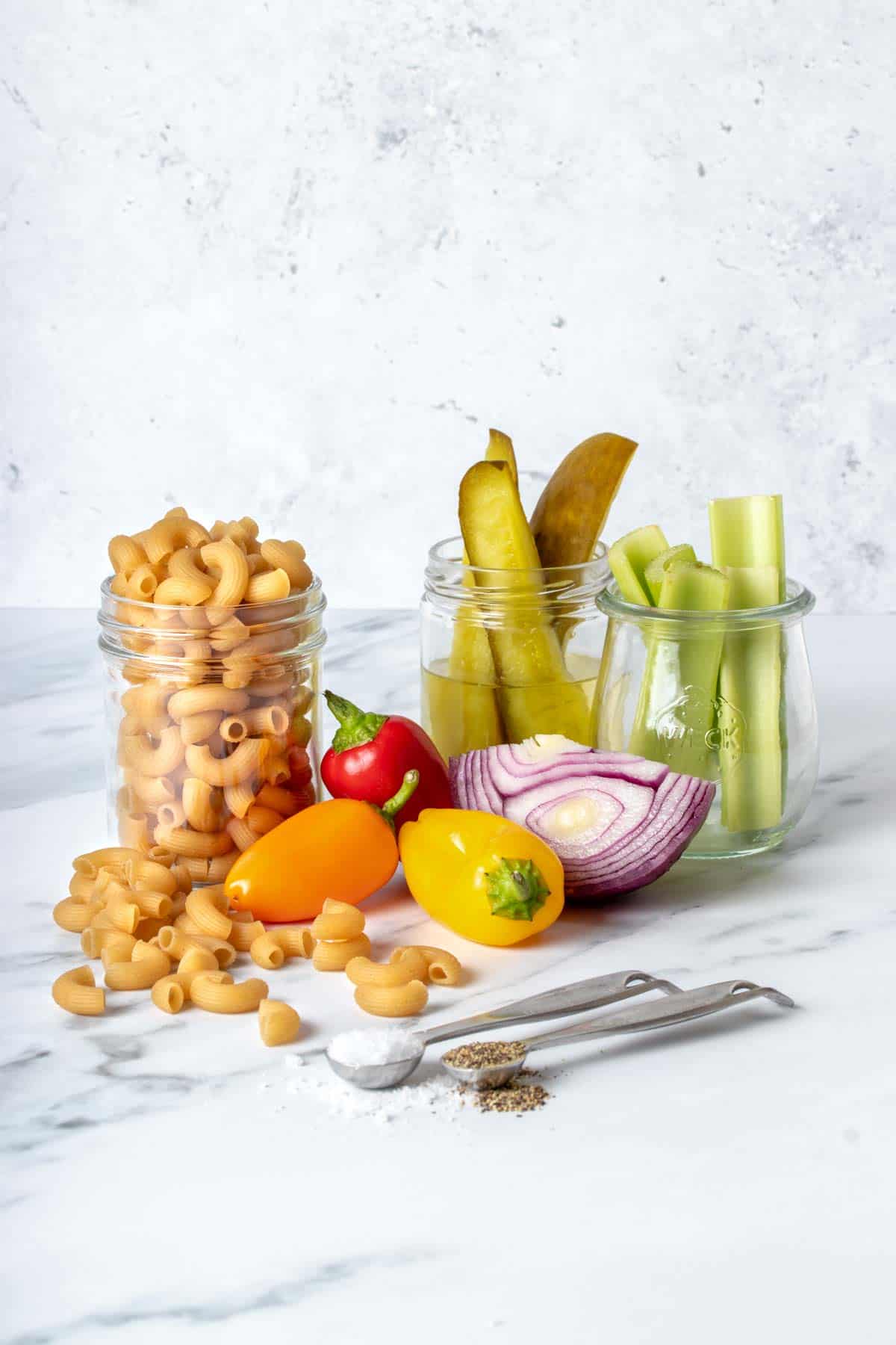 Macaroni, vegetables, pickles and spices in jars and spoons on a marbled surface.