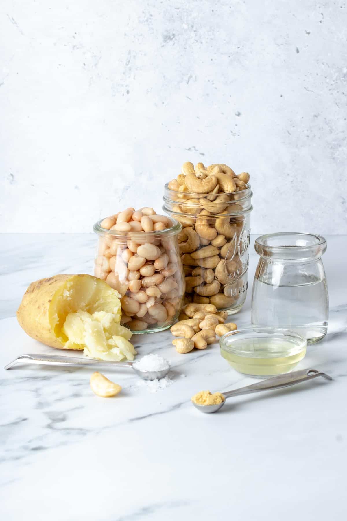 Potato, cashews, white beans, spices and liquids in jars and spoons on a marbled surface.