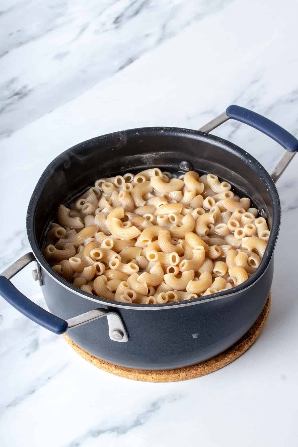 A black pot with handles filled with cooked elbow macaroni on a marbled surface.