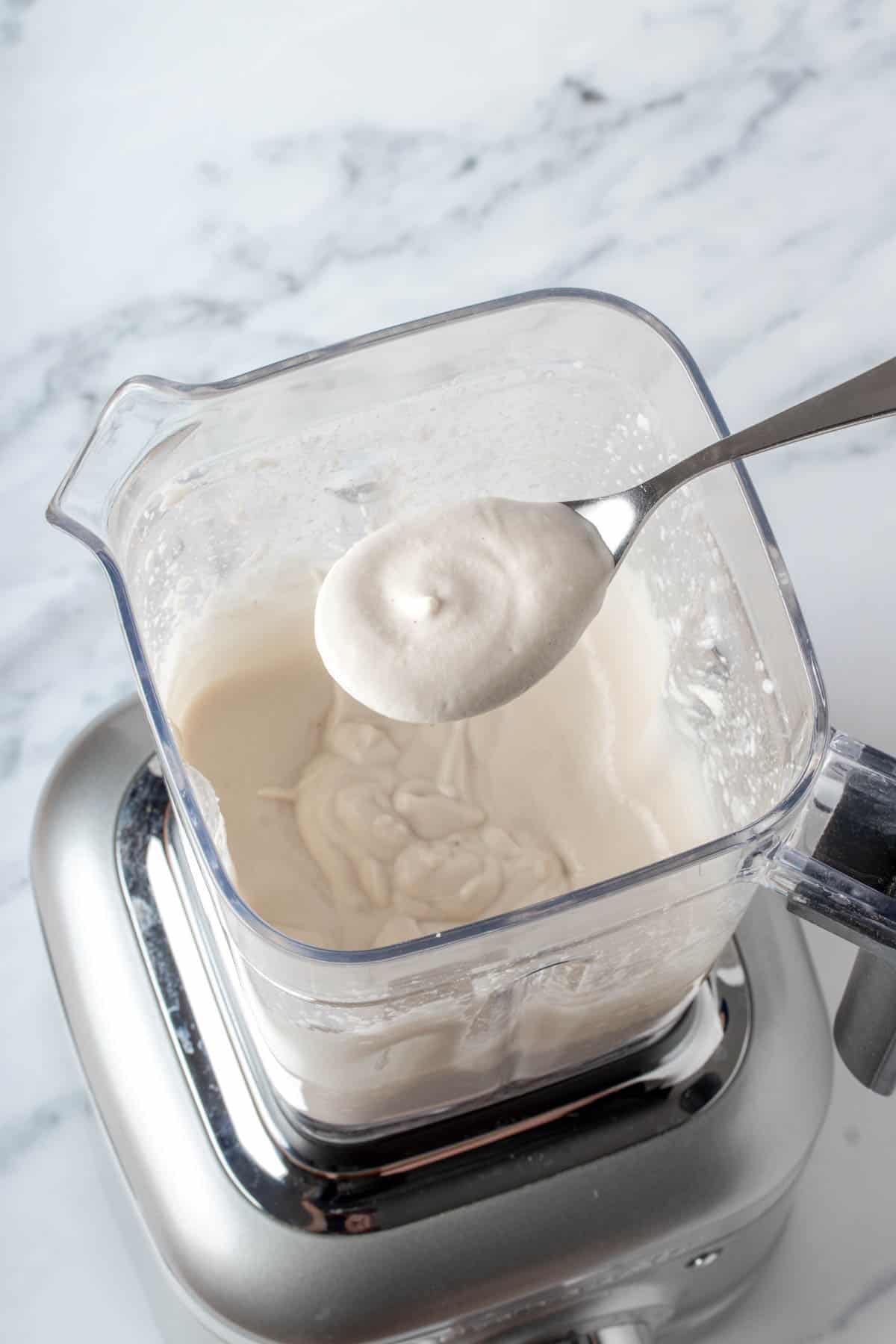 A metal spoon coming out of a silver blender with creamy sauce on it.