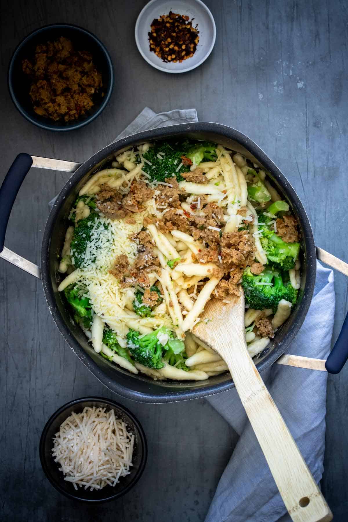 A wooden spoon mixing pasta with broccoli and sausage in a pot