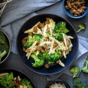 Top view of a dark bowl with bright green broccoli pieces mixed with cavatelli pasta and sausage on a light blue napkin