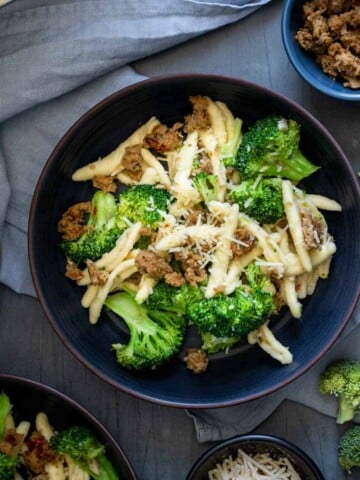 Top view of a dark bowl with bright green broccoli pieces mixed with cavatelli pasta and sausage on a light blue napkin