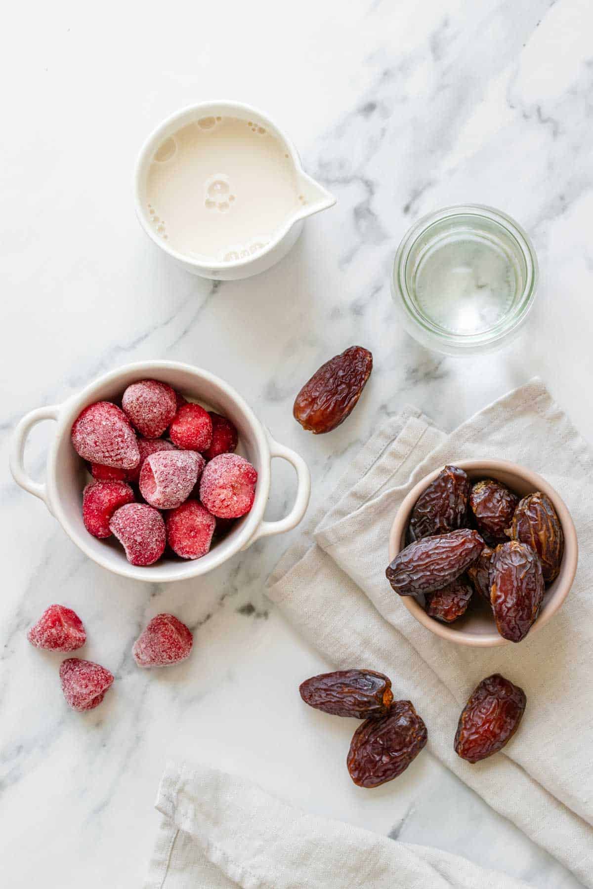 Top view of a marble surface with bowls of water, dates, milk and frozen strawberries 