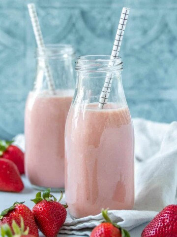 Pink strawberry milk in two glass jars with straws sitting in front of each other around strawberries