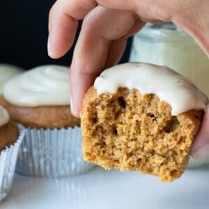 A pumpkin cupcake with white frosting and a bite out of the front being held up by a hand.