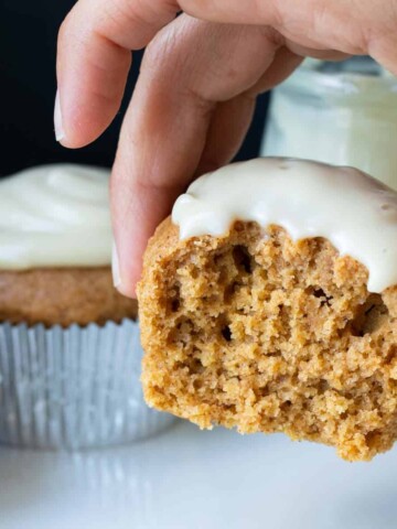 A pumpkin cupcake with white frosting and a bite out of the front being held up by a hand.
