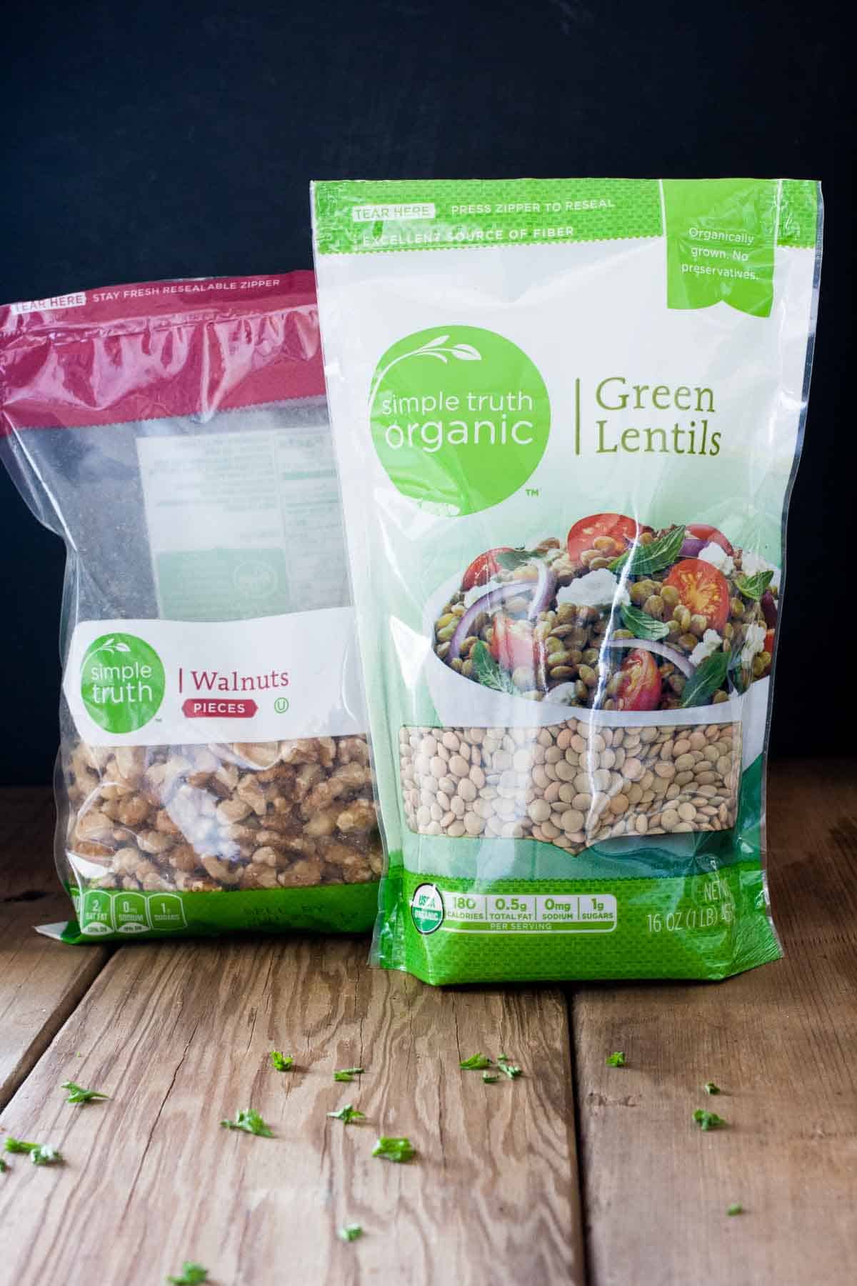 A bag bag of walnuts with a pink top next to a bag of lentils with green edges on a wooden surface.