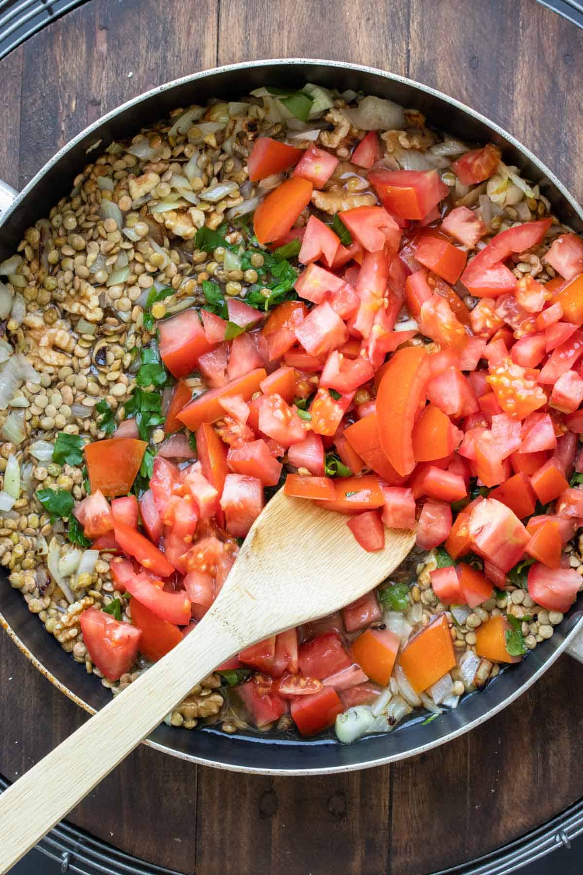 A wooden spoon mixing chopped tomatoes in a pan with lentils and parsley.