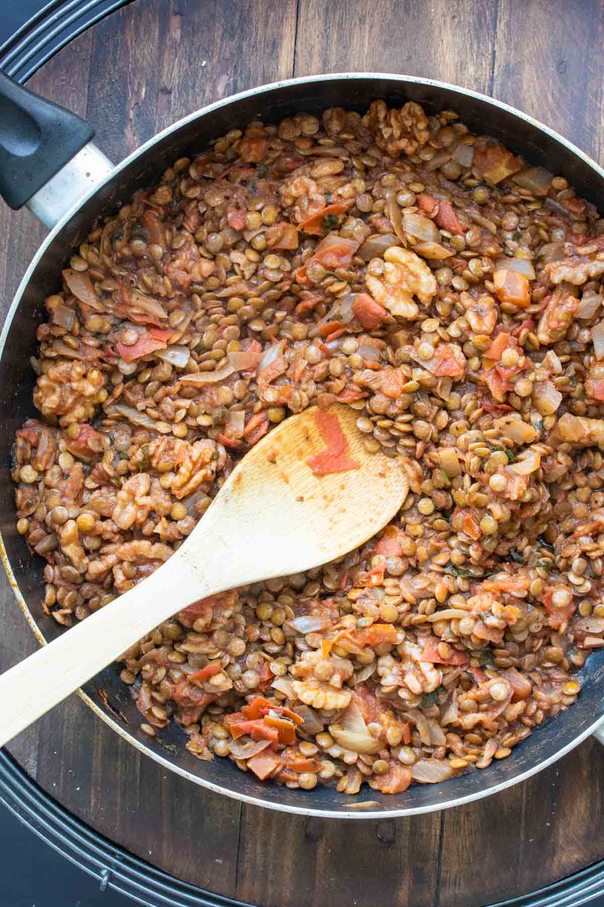 A wooden spoon mixing a mixture of lentils and tomatoes in a pan.