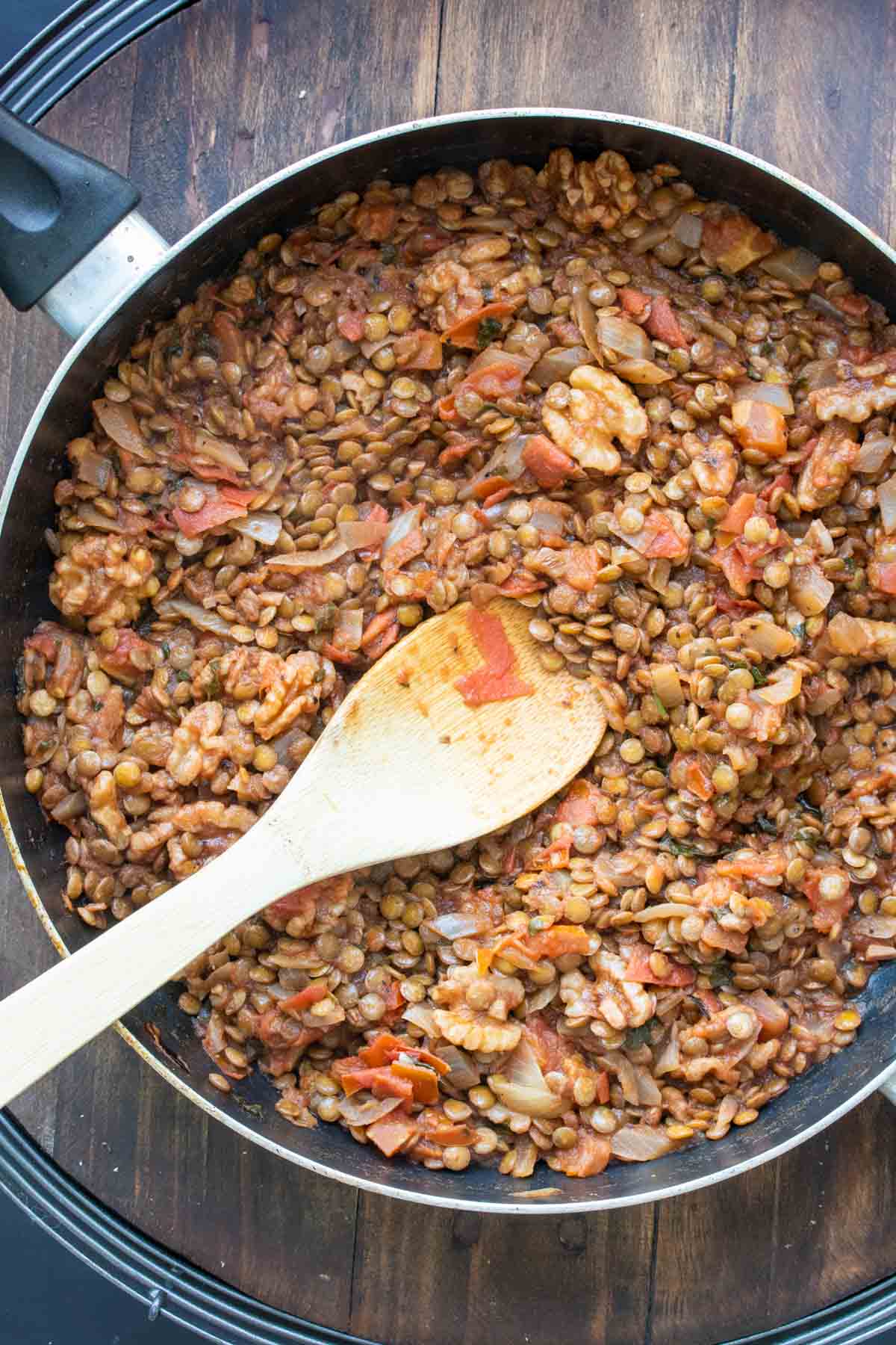 A wooden spoon mixing a mixture of lentils and tomatoes in a pan.