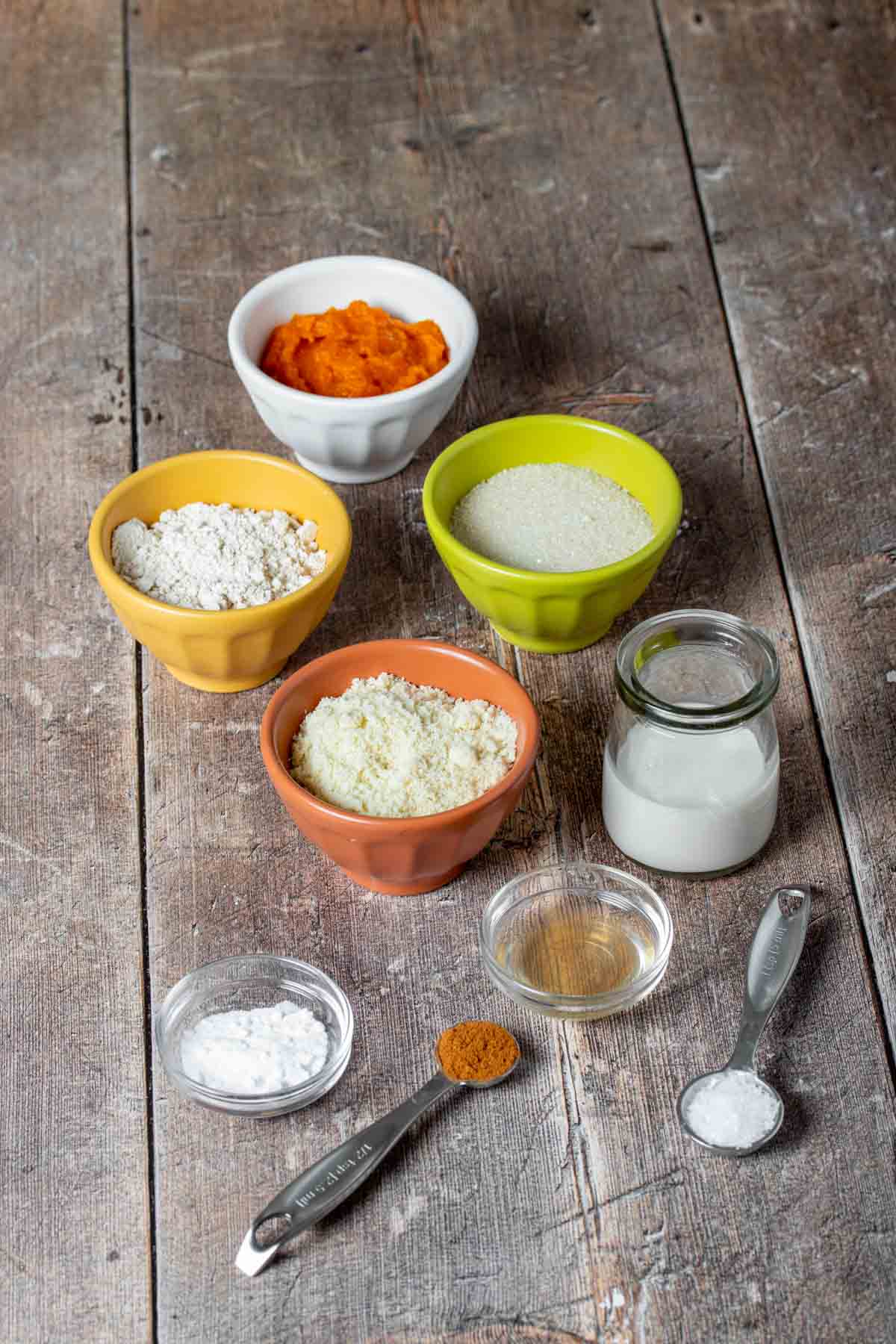 A wooden table with flours, pumpkin puree, milk, vanilla extract and spices in bowls and spoons.