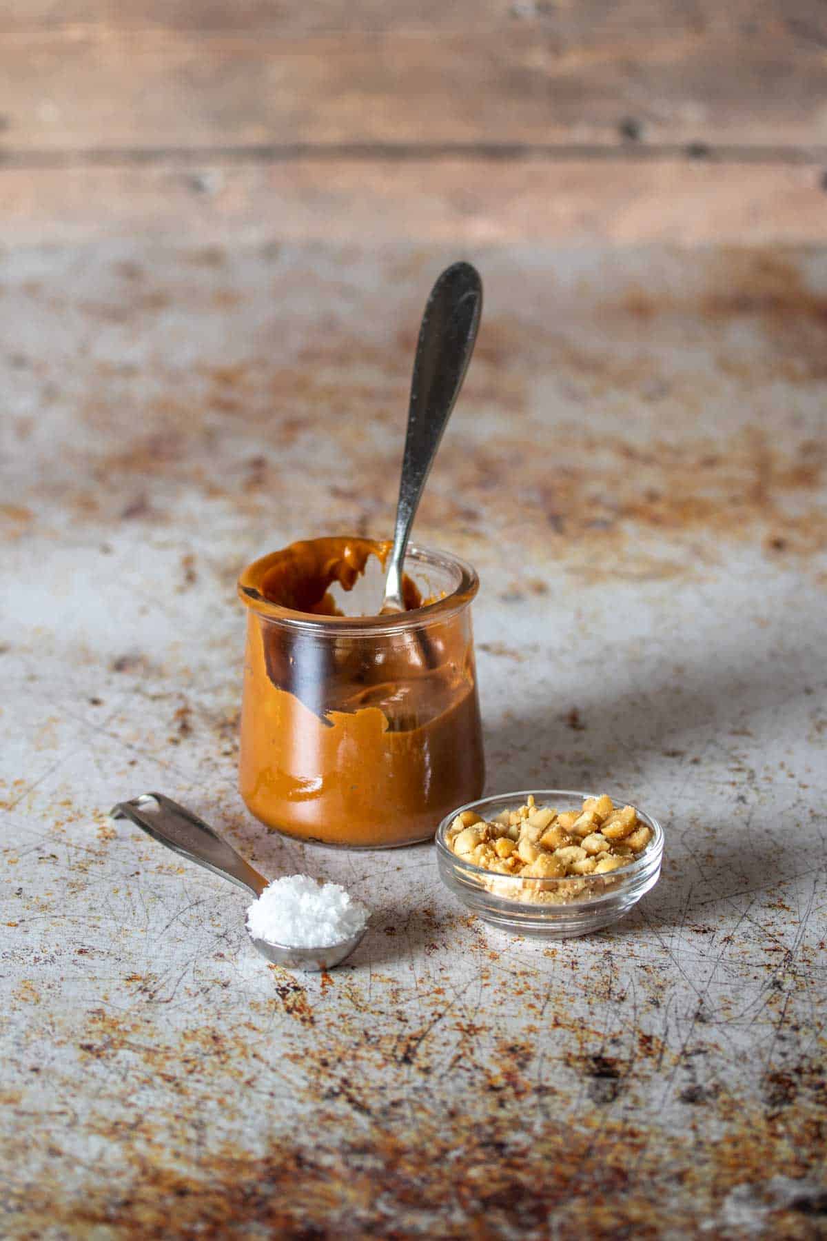 A jar of caramel, spoon of flaked sea salt and glass bowl of chopped peanuts on a speckled tan and white surface.