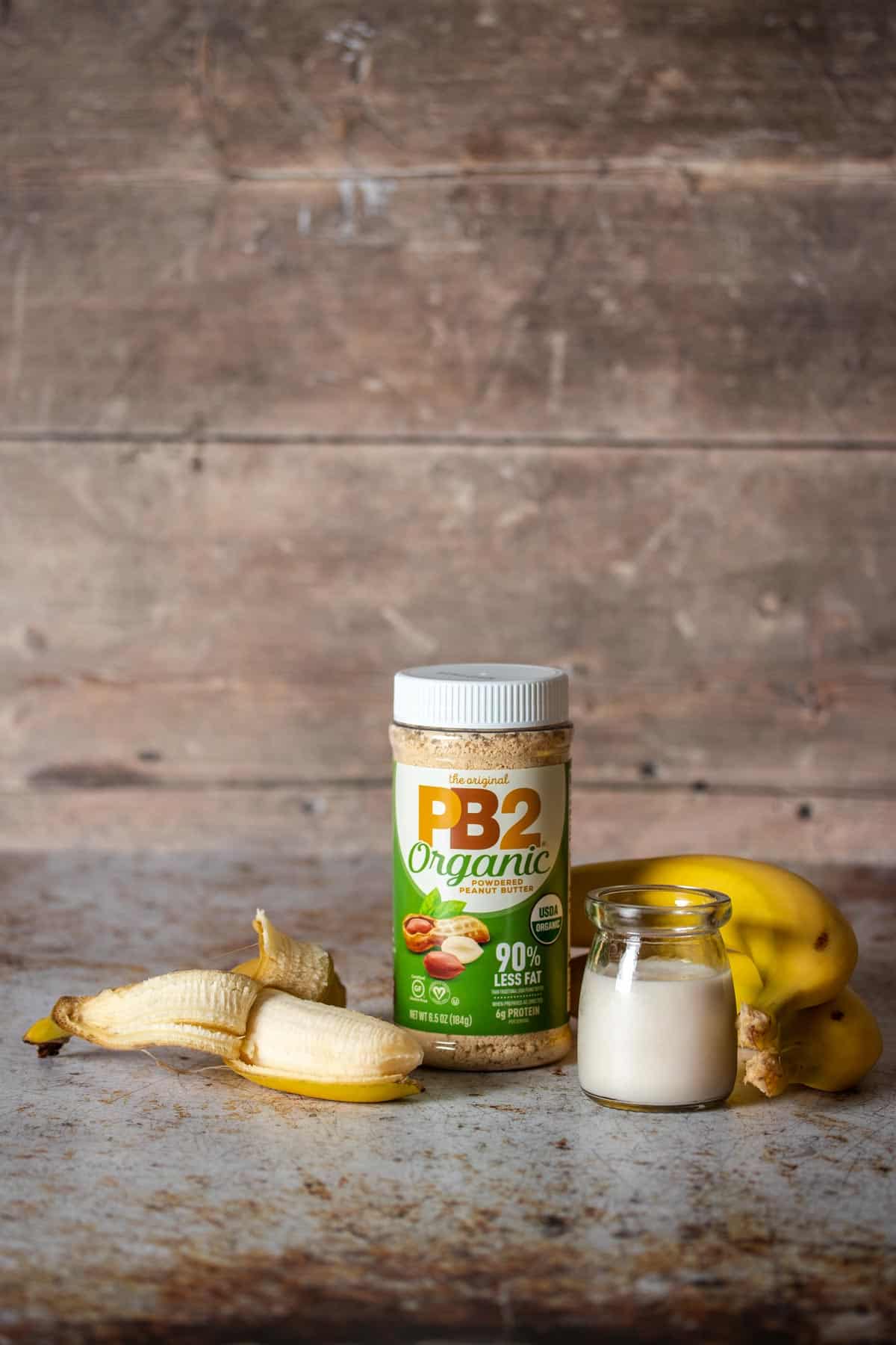 An open banana, closed bananas, jar of milk and container of powdered peanut butter with a green and white label.