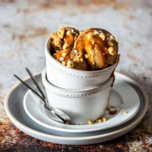 Scoops of a tan ice cream topped with caramel and peanuts in two stacked white bowls on stacked white plates.