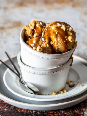 Scoops of a tan ice cream topped with caramel and peanuts in two stacked white bowls on stacked white plates.
