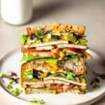 Two halves of a veggie and tofu sandwich stacked on top of each other on a tan plate in front of a glass of milk.