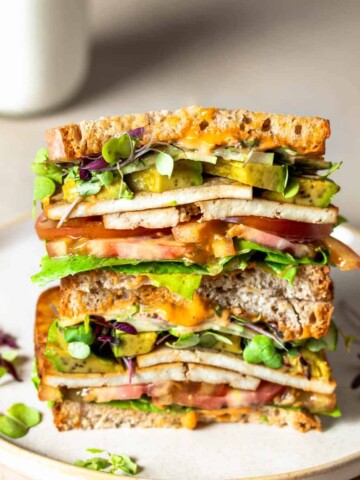 Two halves of a veggie and tofu sandwich stacked on top of each other on a tan plate in front of a glass of milk.