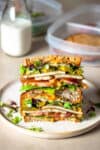 A stack of two halves of a veggie and tofu sandwich stacked on top of each other on a plate in front of sectioned plastic containers and a glass of milk.