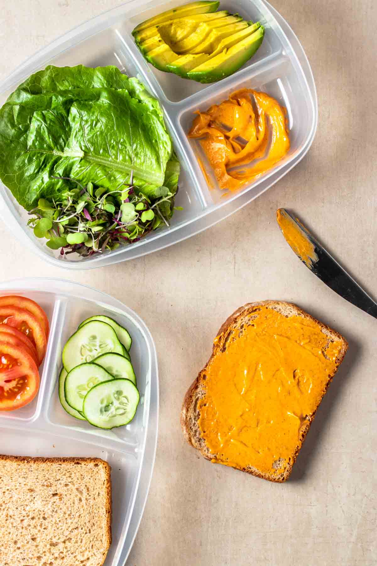 Top view of plastic containers with sections filled with veggies and sliced bread and a piece of bread with sauce on it next to a knife.