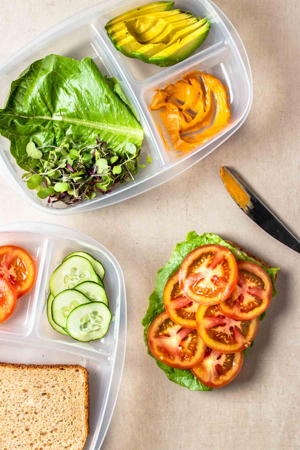 A piece of bread with lettuce and tomatoes on top next to a knife and containers with sandwich toppings in them.