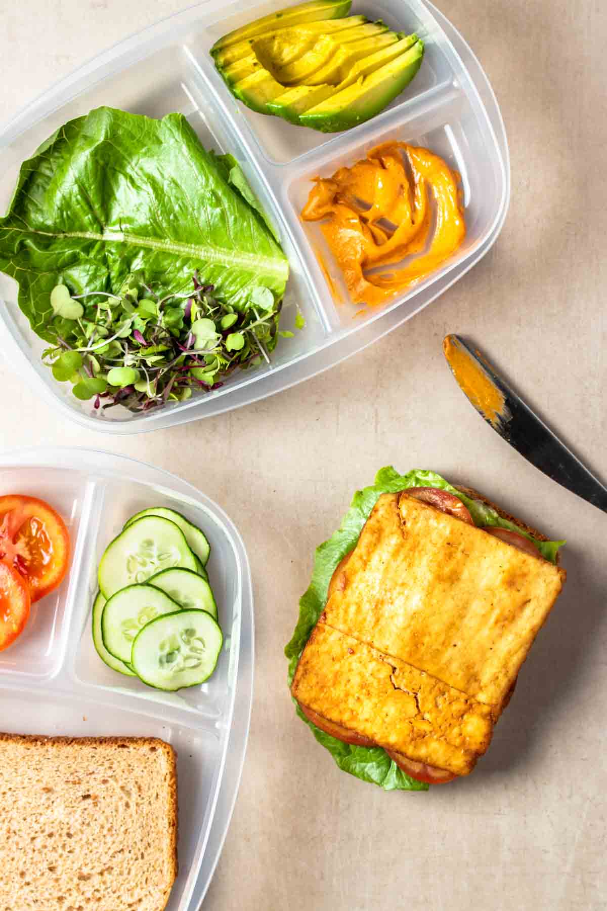 A slice of baked tofu on a piece of bread with tomatoes and lettuce under the tofu next to sectioned containers with more toppings and a knife.