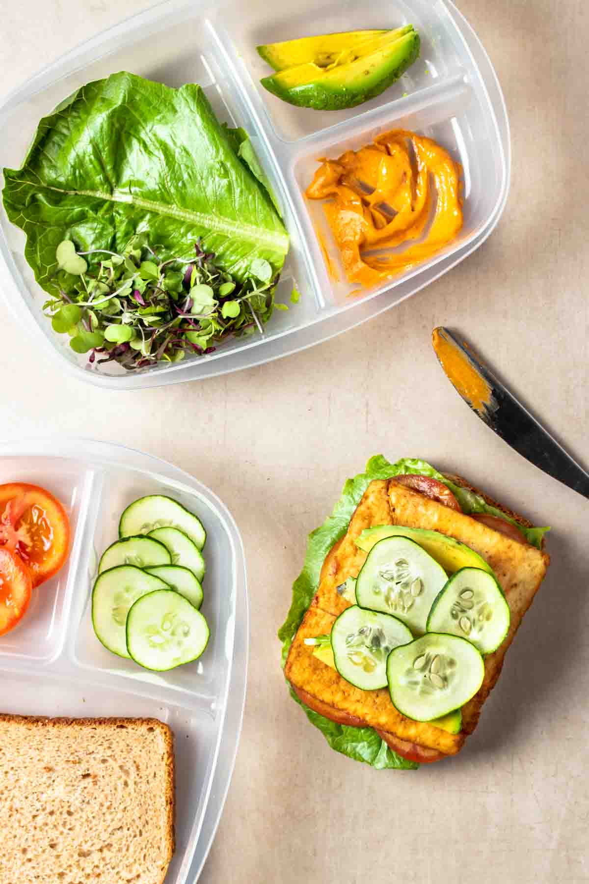 Sectioned containers with sandwich toppings next to a knife and an open faced sandwich with lettuce, sliced tofu and cucumbers on top.