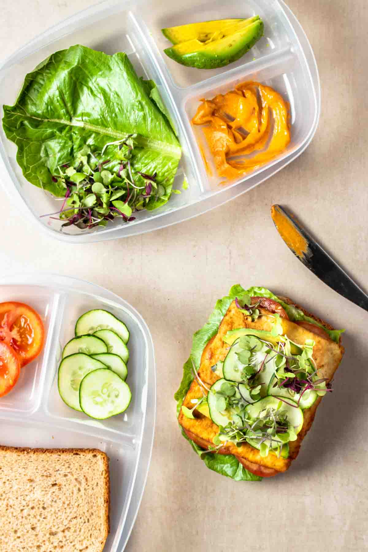 Containers with sections and sandwich toppings in them next to a sandwich being build with lettuce, tofu, cucumbers and sprouts on top next to a knife.
