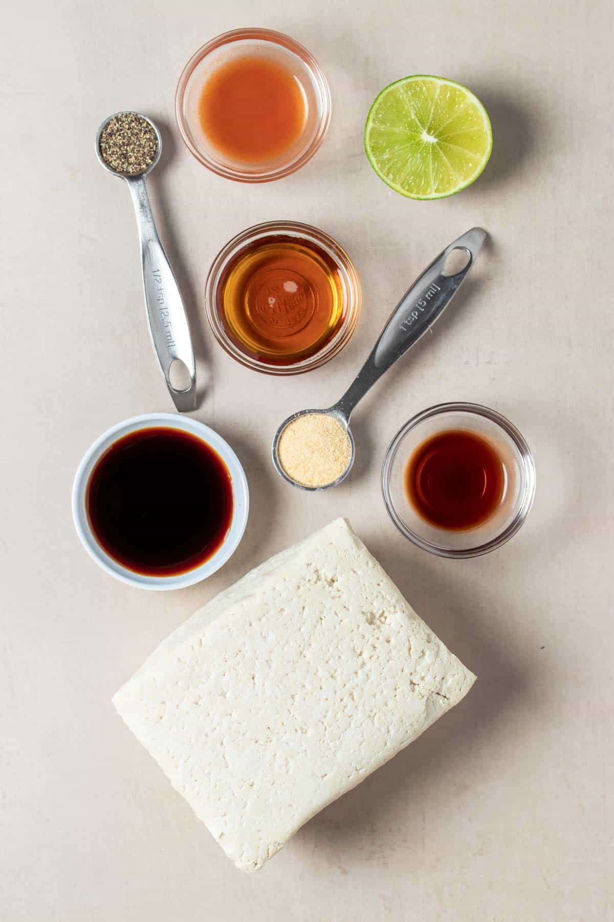 Top view of a block of tofu next to glass bowls of brown liquids, a lime half and measuring spoons with spices.