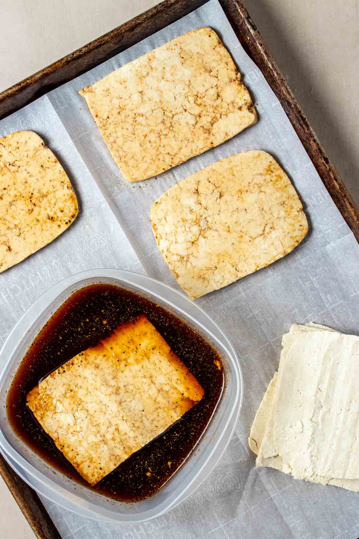 Top view of a parchment lined baking sheet with sliced tofu marinating in a container with brown liquid and some slices on the parchment.