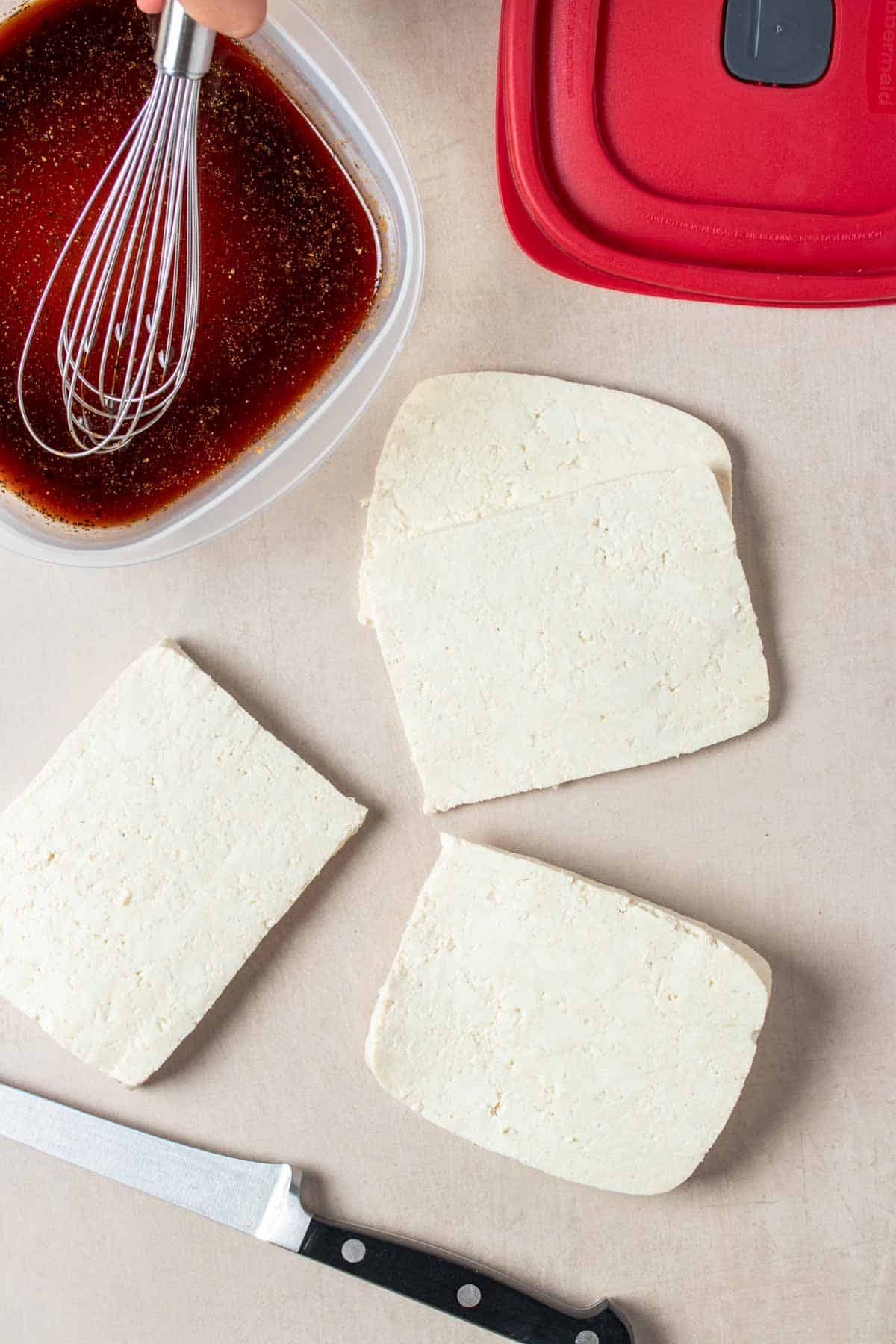 Top view of thinly sliced tofu and brown liquid in a plastic container being whisked next to a red lid.