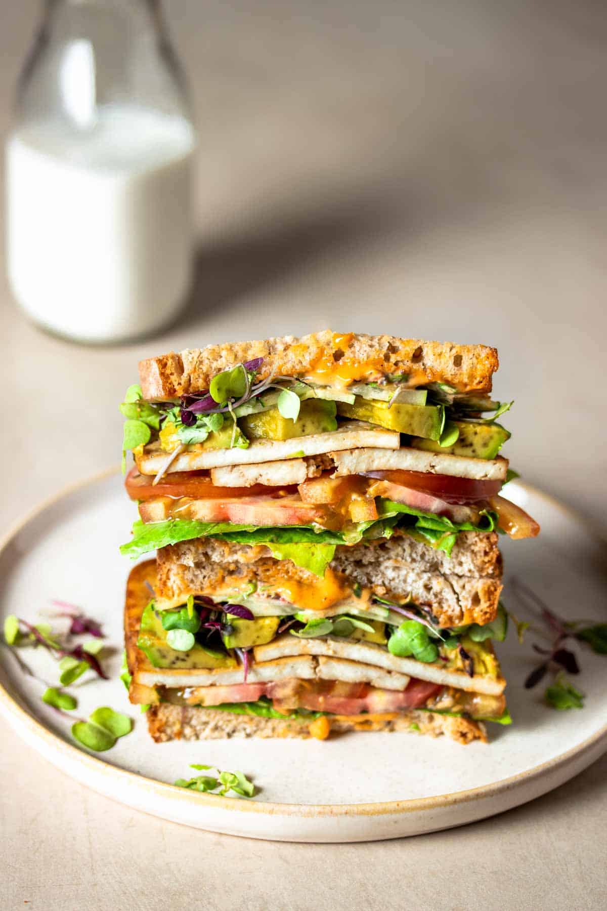 A tofu and veggie sandwich that has been cut in half and stacked on a tanish plate in front of a glass of milk.