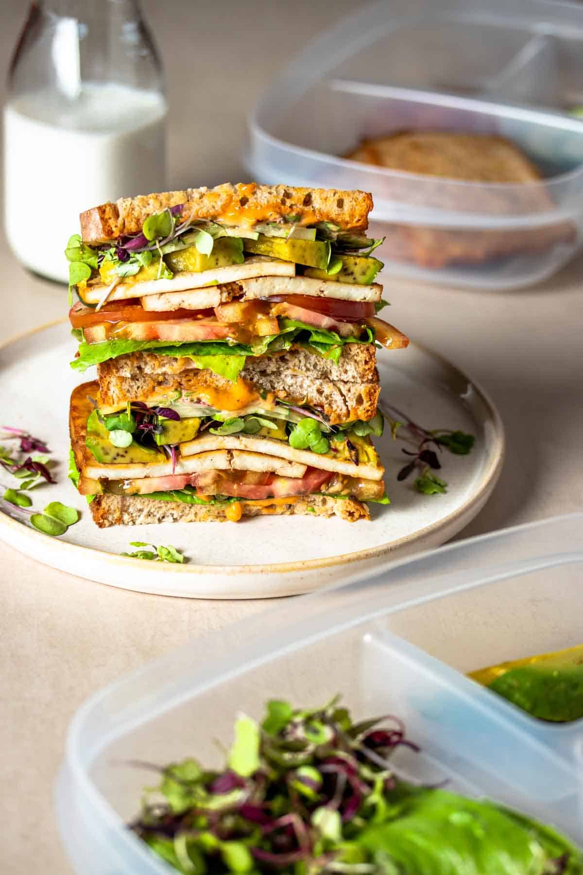A plate with two halves of a tofu veggie sandwich stacked on each other surrounded by plastic containers and a glass of milk.
