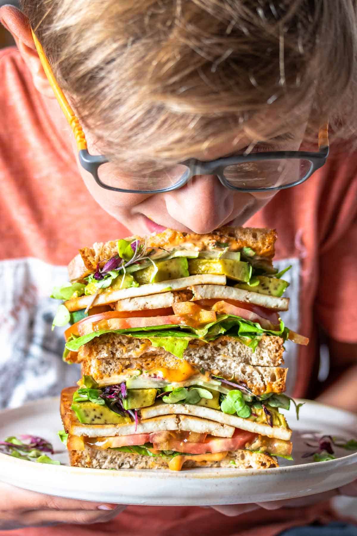 A boy with glasses and an orange shirt leaning down and taking a bit from the top of a stack of tofu and veggie sandwich halves.
