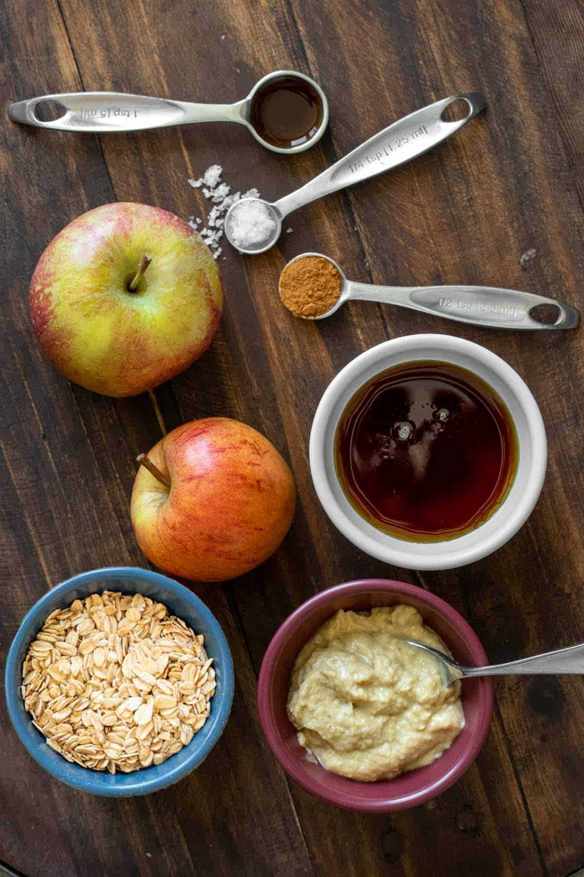 Bowls of oats, cashew butter and maple syrup next to apples and spoons of spices.